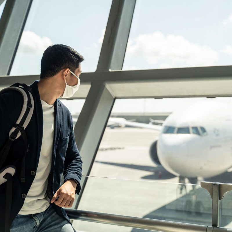 Asian traveler business man wearing face mask waiting to board into airplane, standing in departure terminal in airport. Male passenger traveling by plane transportation during covid19 virus pandemic.